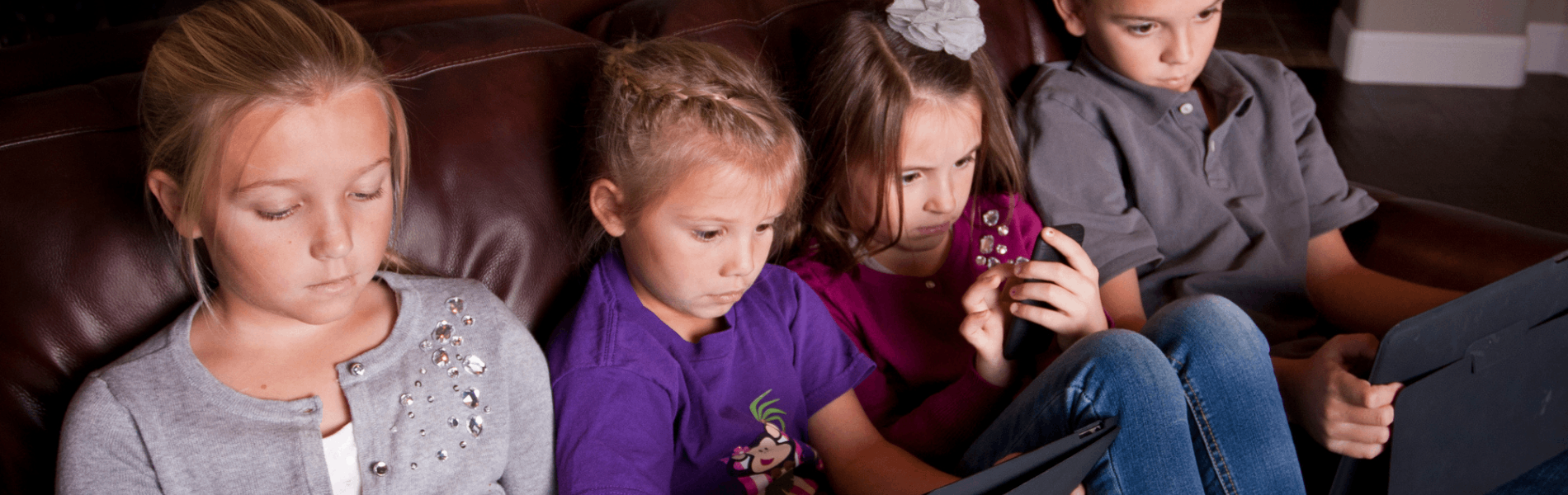 A row of four young children sitting on a leather couch, fixated on the mobile devices they are each holding.